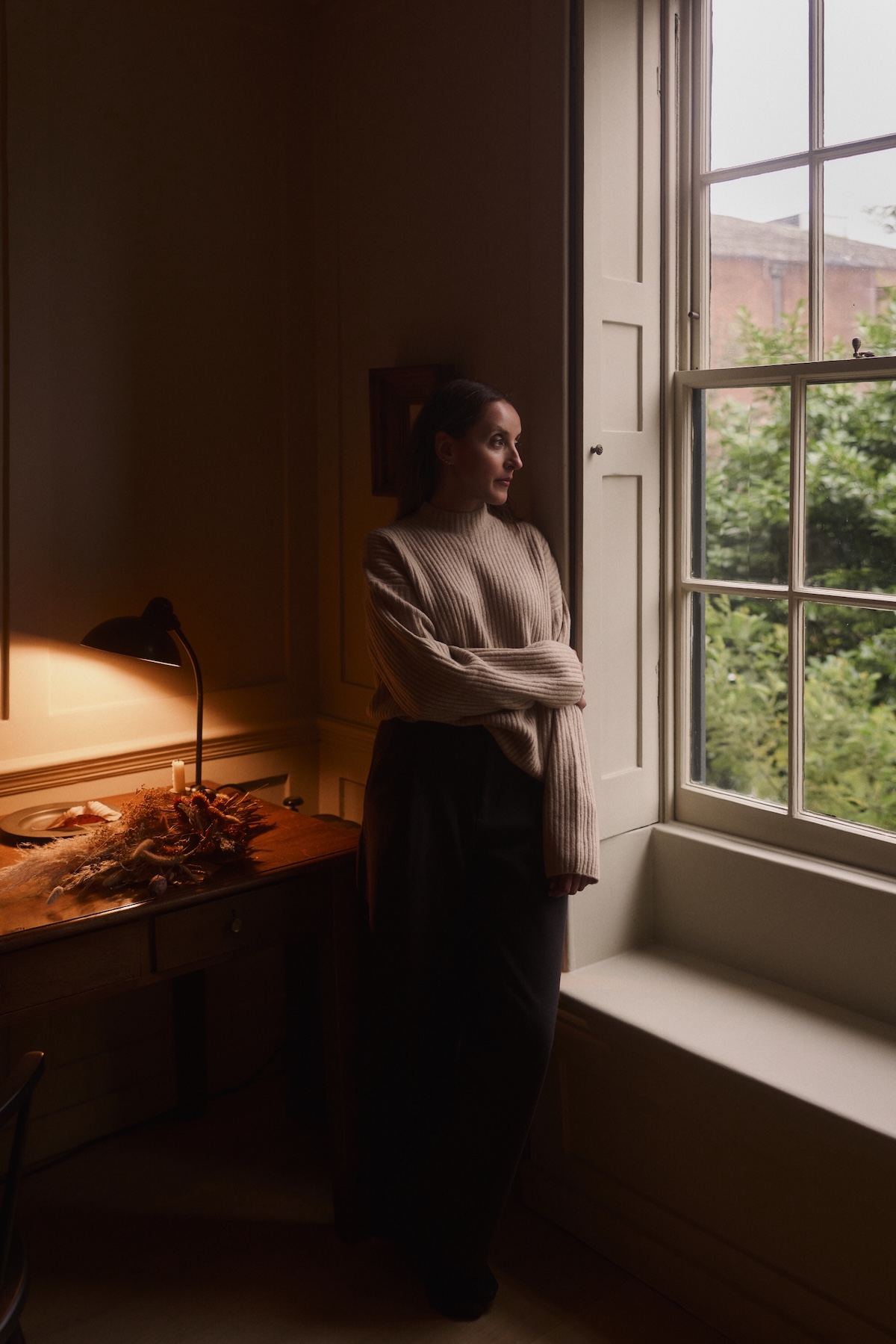 A woman in a cozy sweater stands by a large window, looking outside thoughtfully. Warm tones and soft lighting create a serene, reflective mood.
