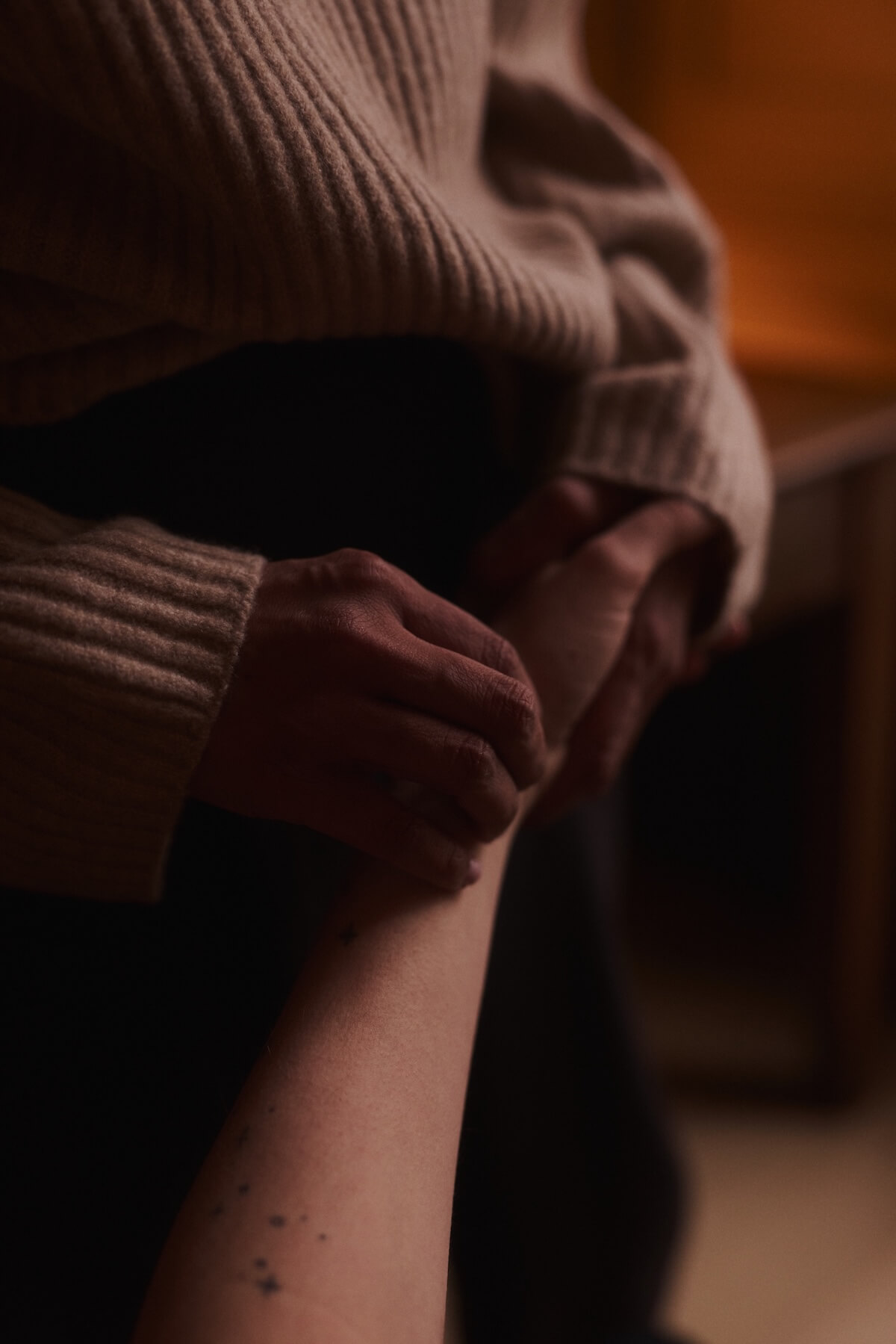 A person in a beige sweater gently holds clinets's wrist in a dimly lit room, conveying warmth and care.