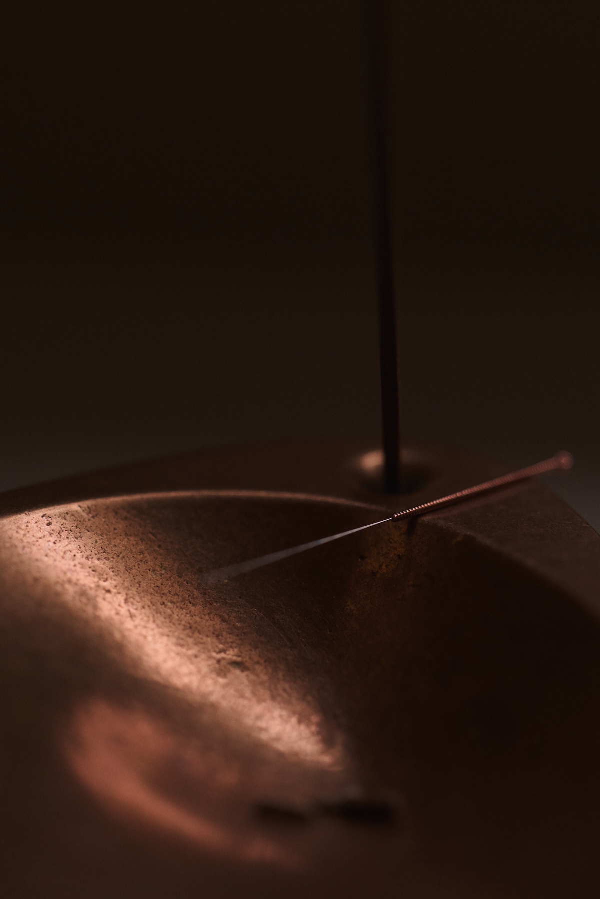 A fine acupucture needle balancing on the edge of a copper bowl, set in against a dark, calm background, conveying precision and care.