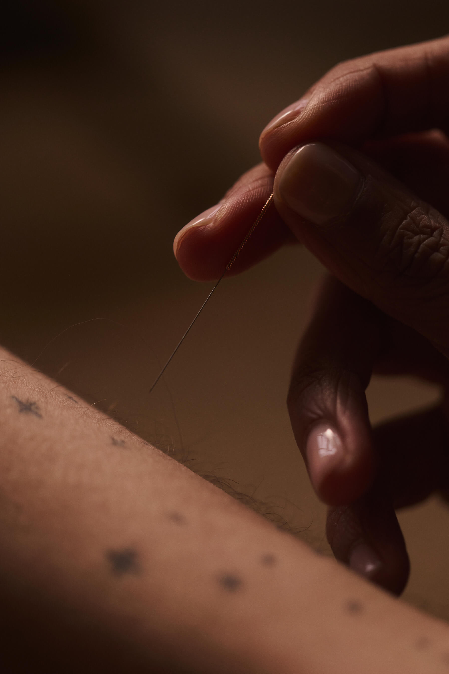Close-up of an acupucture needle in focus, near a forearm with faint star tattoos. Dim, warm lighting.