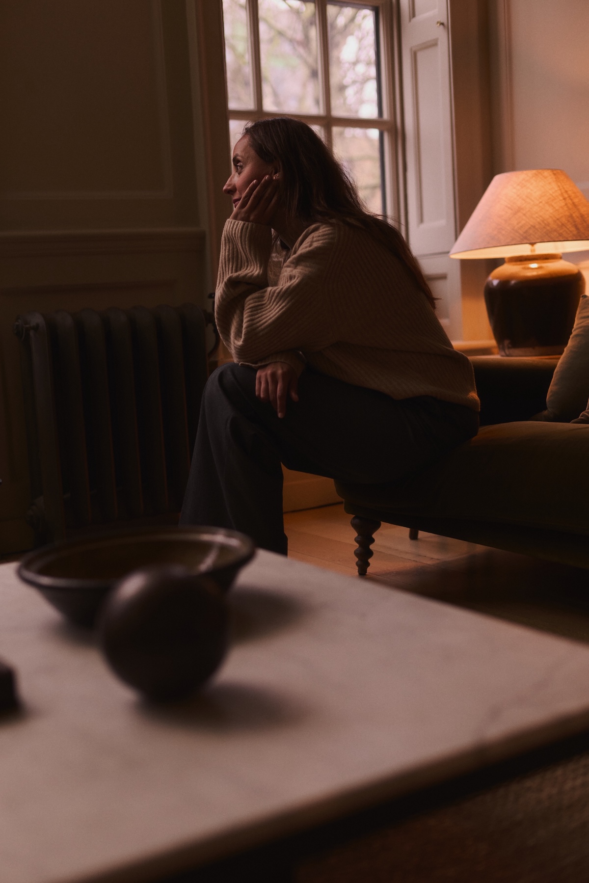 Asha Chong, acupuncturist sitting calmly on the edge of a sofa in a dimly light room. A warm lamp is on in the background, showing a relaxed and calm environment.